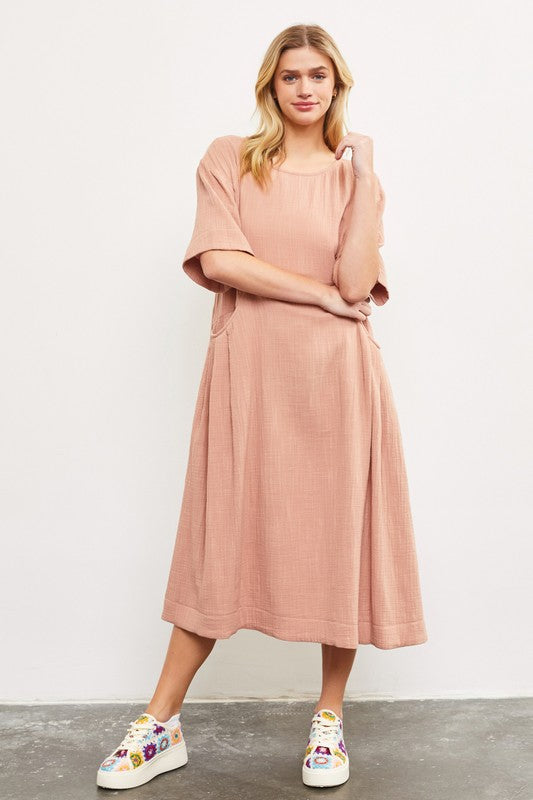 Woman wearing a blush pink cotton midi dress with side pockets.